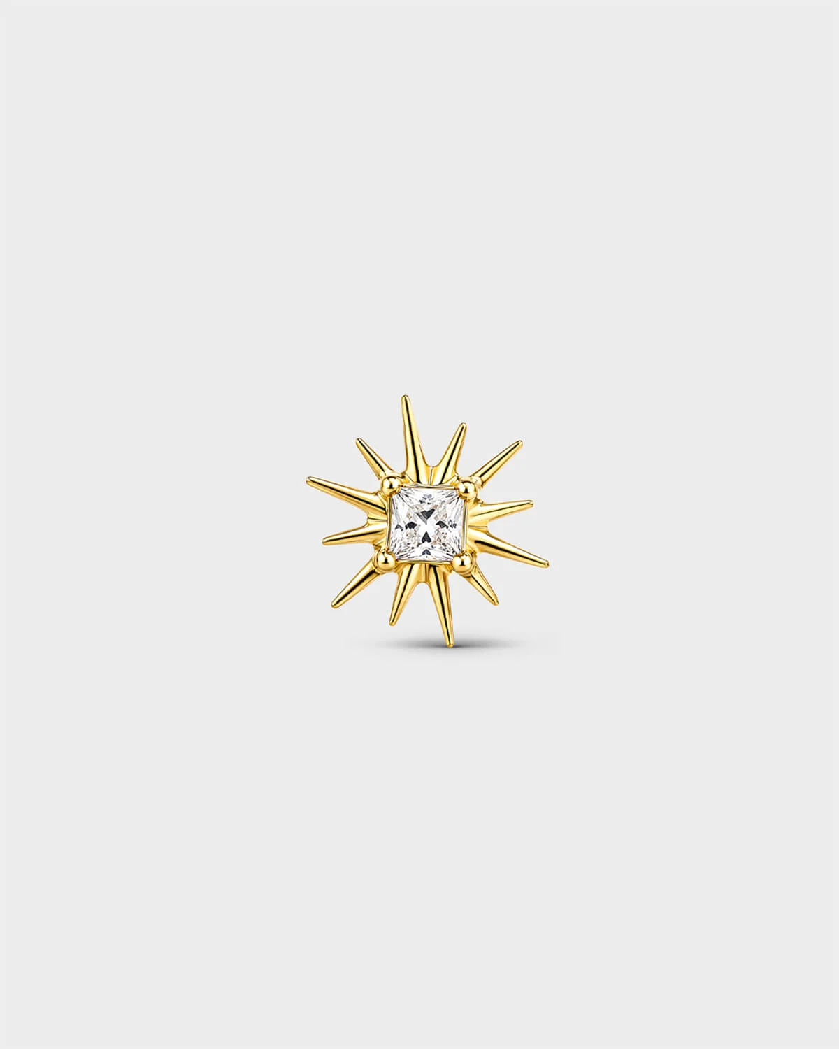 Gold titanium threadless end with a princess-cut clear stone set within a sculpted starburst design. On a light grey background.