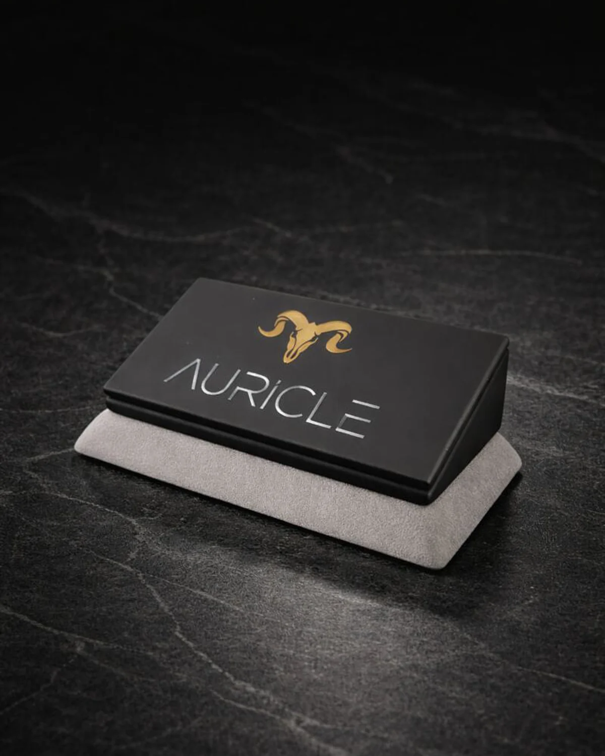 AURICLE jewellery branded nameplate stand. The stand has a light gray suede base with a black leather top. Sat on a dark worktop with moody lighting.