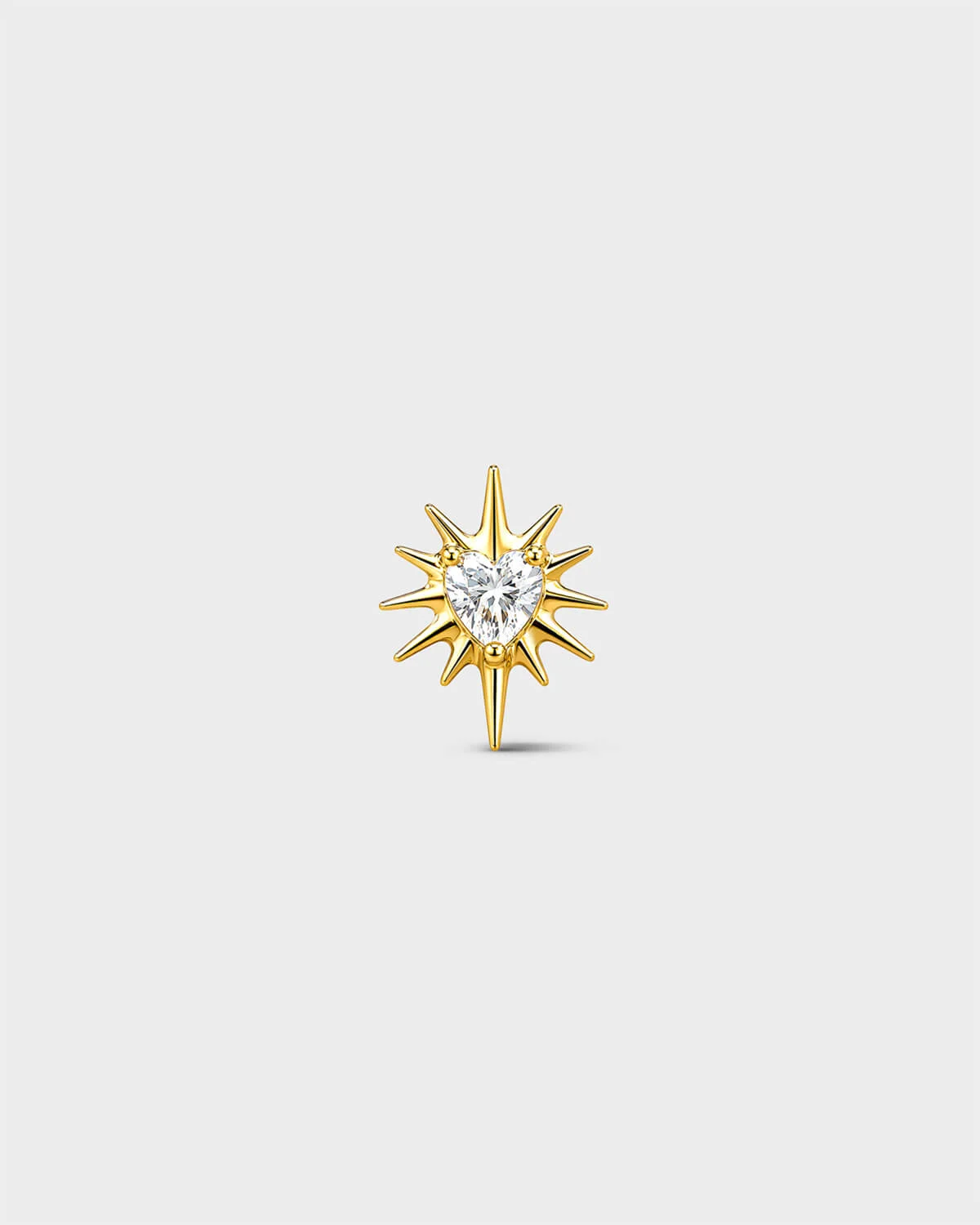 Gold titanium threadless end earring with a heart-cut clear stone set within a sculpted starburst design. On a light grey background.